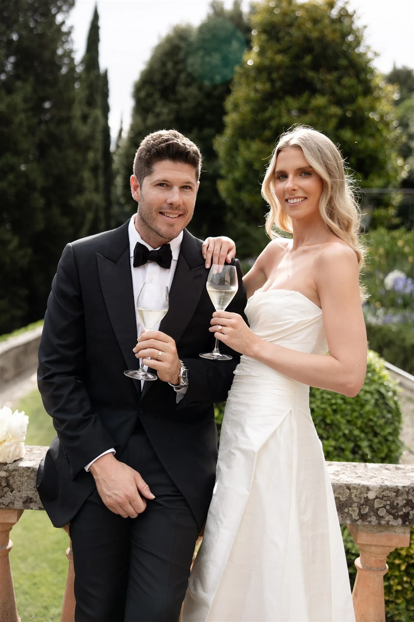Couple in wedding attire holding wine glasses beside a stone balustrade