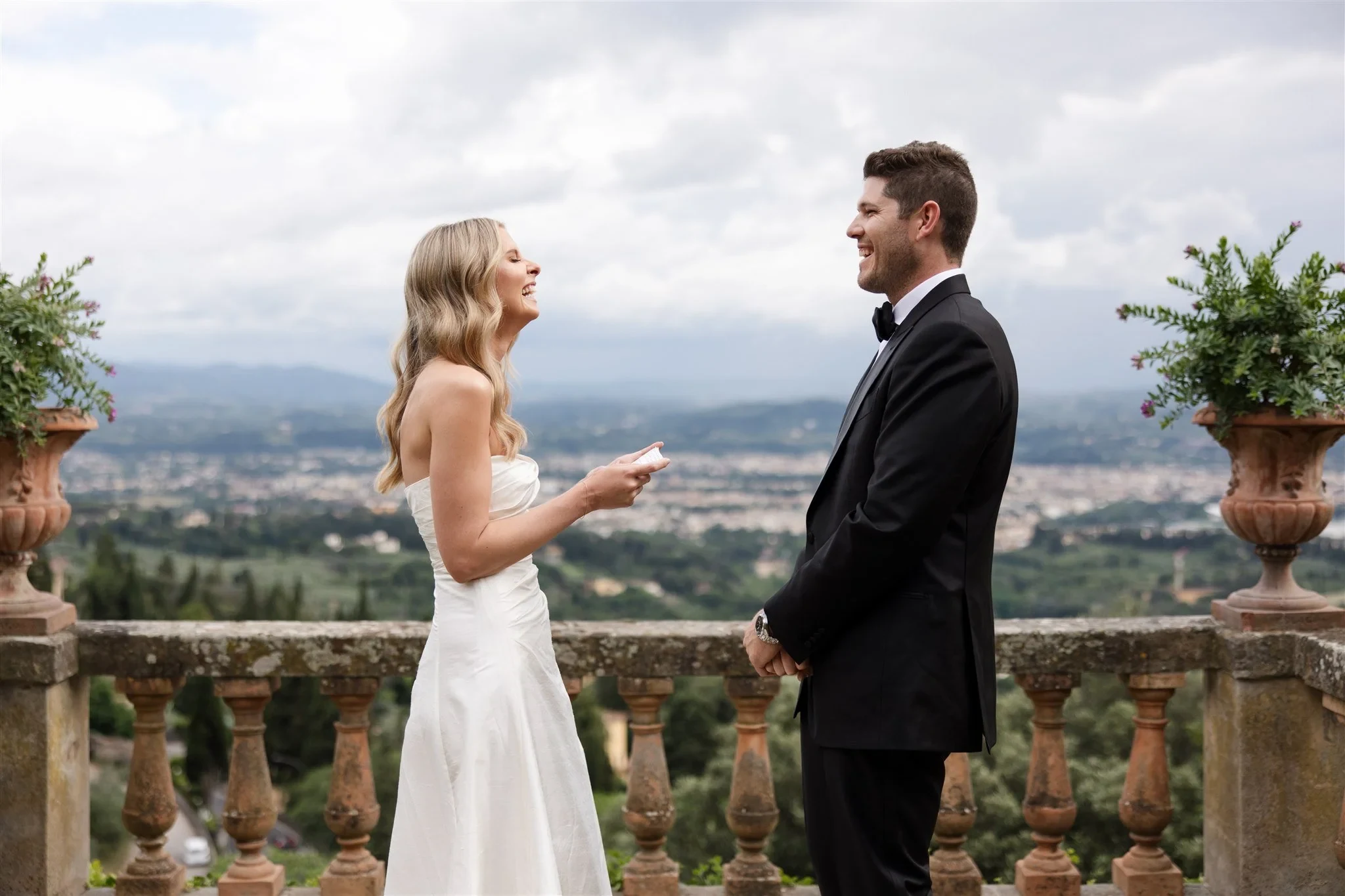Holly smiling at Matt with her vow card in hand and Florence in the distance, at Belmond Villa San Michele in Fiesole, overlooking Florence, Tuscany, Italy, for an Elopement Collective elopement. Photo by Khara from House of Lucie.