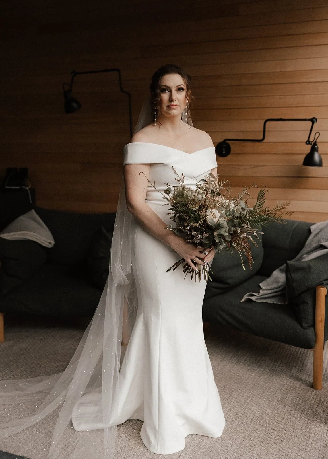 Sarah holding her bouquet by The Romantics in front of a pale timber wall and green armchair, at Pumphouse Point, Lake St Clair, Tasmania, for an Elopement Collective elopement. Photo by Bulb Creative.