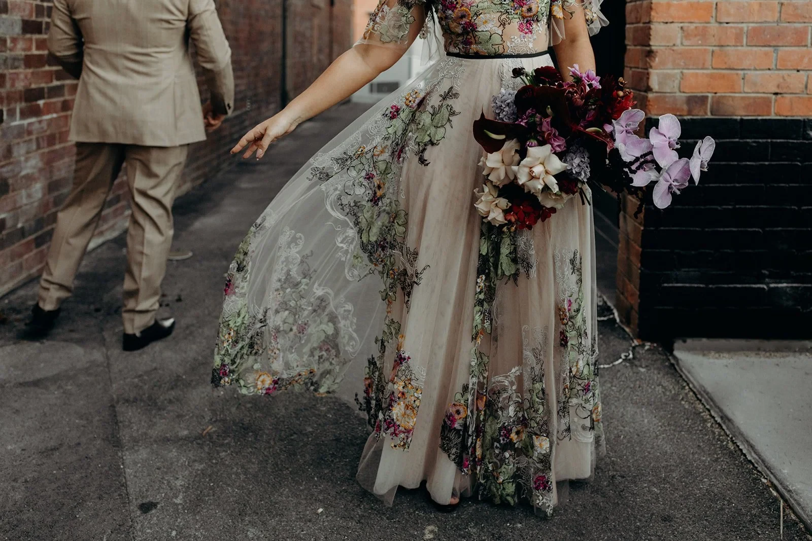 Victoria lifting the sheer floral layer of her skirt while holding her bouquet by Mrs Gibbons Flowers, with Daniel walking ahead, in Fortitude Valley, Brisbane, for an Elopement Collective elopement. Photo by Tessa Shannon.