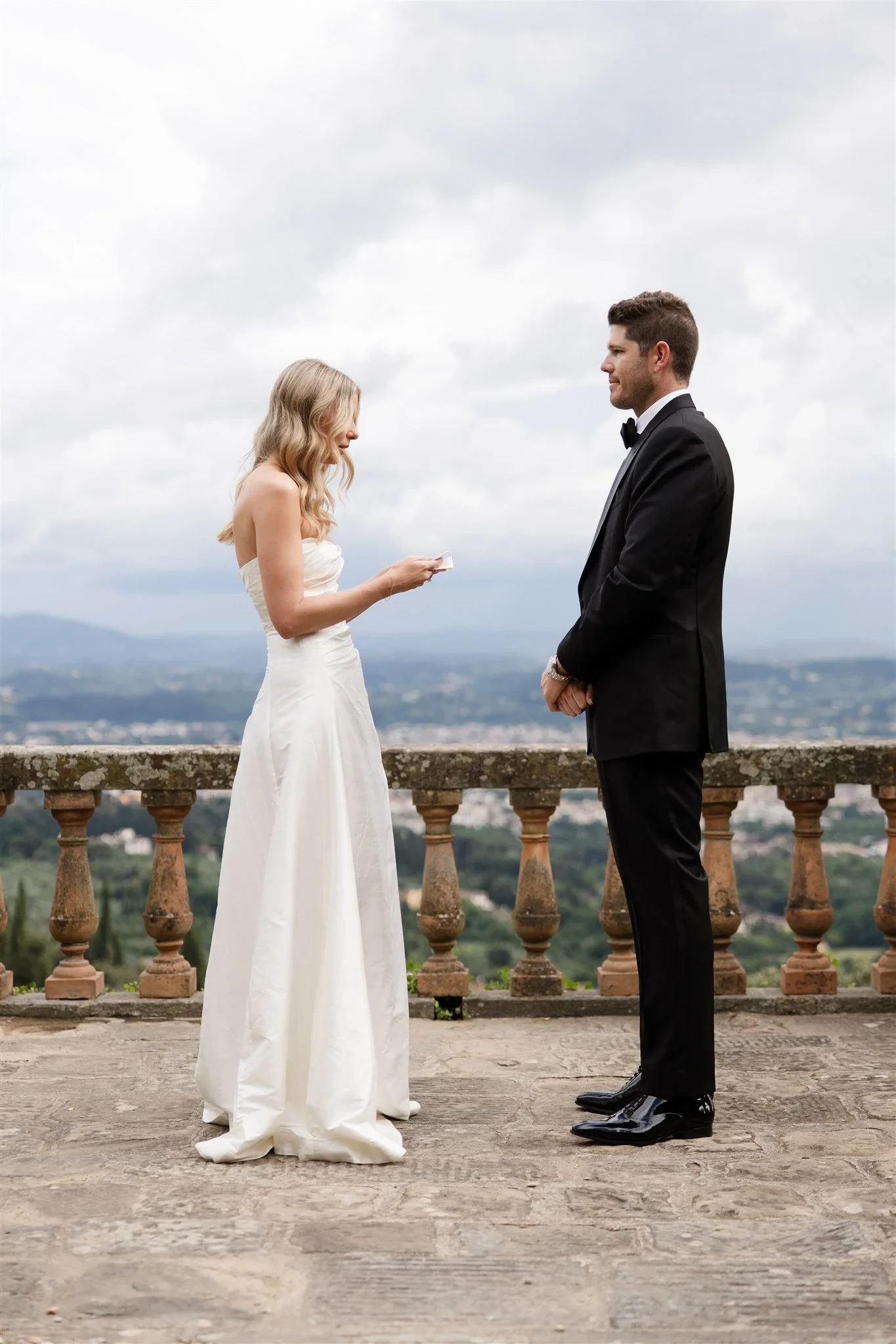 Holly reading from a vow card as Matt faces her beside a stone balustrade overlooking Florence, at Belmond Villa San Michele in Fiesole, overlooking Florence, Tuscany, Italy, for an Elopement Collective elopement. Photo by Khara from House of Lucie.