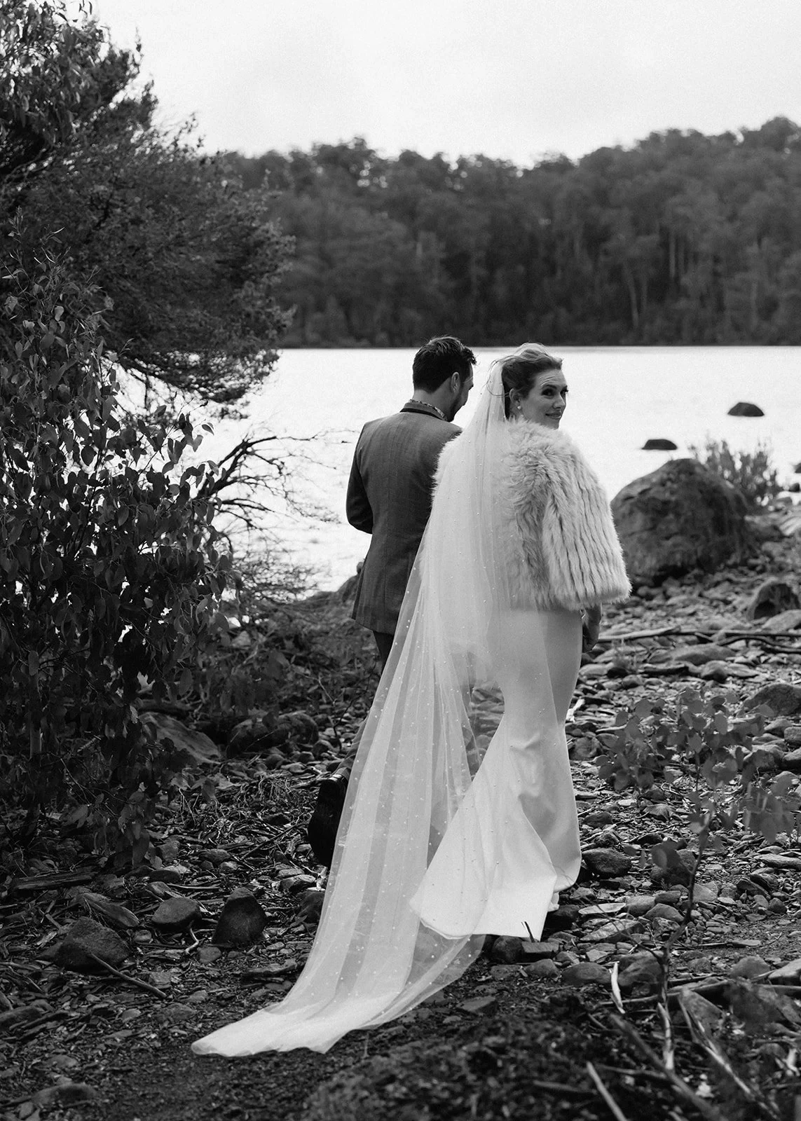 Black-and-white portrait of Sarah looking back over her shoulder while walking beside Jono on the lakeshore, at Pumphouse Point, Lake St Clair, Tasmania, for an Elopement Collective elopement. Photo by Bulb Creative.