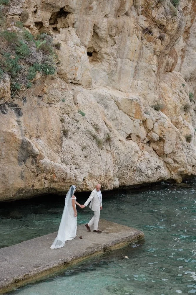 A peek into eloping in Sicily with The Elopement Collective. A couple is walking away from a stone wall into water at sunset with an overexposed sky and light haze.