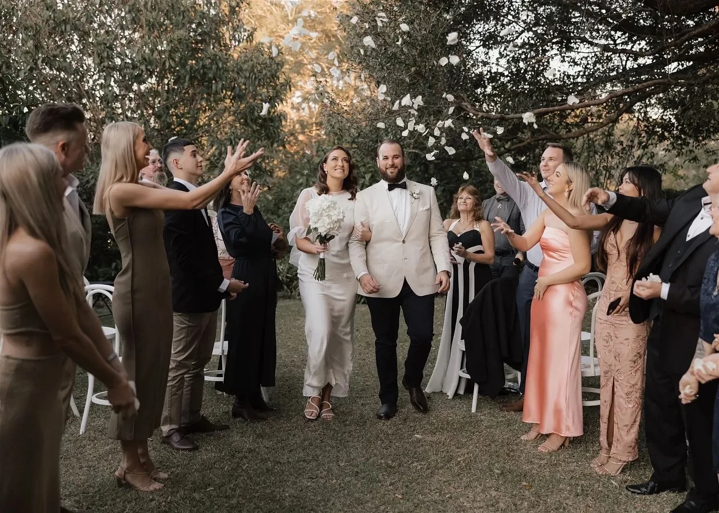 Tineeka and Chris walking between cheering guests beneath garden trees, her bouquet by Fifth Ave in hand, in Montville, Sunshine Coast Hinterland, Queensland, for an Elopement Collective elopement. Photo by Bulb Creative.