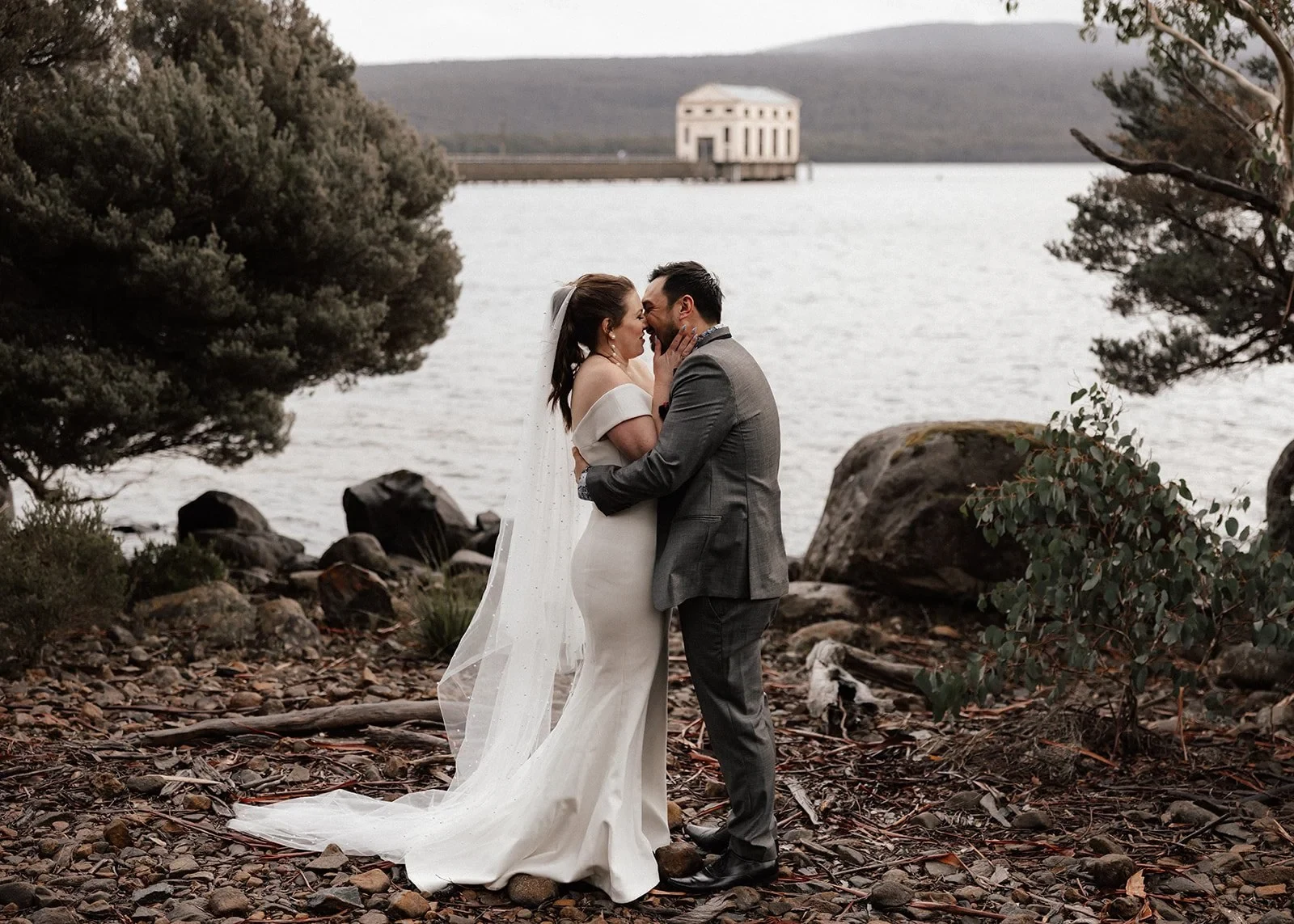 Sarah and Jono kissing on the rocky lakeshore with the Pumphouse building behind them, at Pumphouse Point, Lake St Clair, Tasmania, for an Elopement Collective elopement. Photo by Bulb Creative.