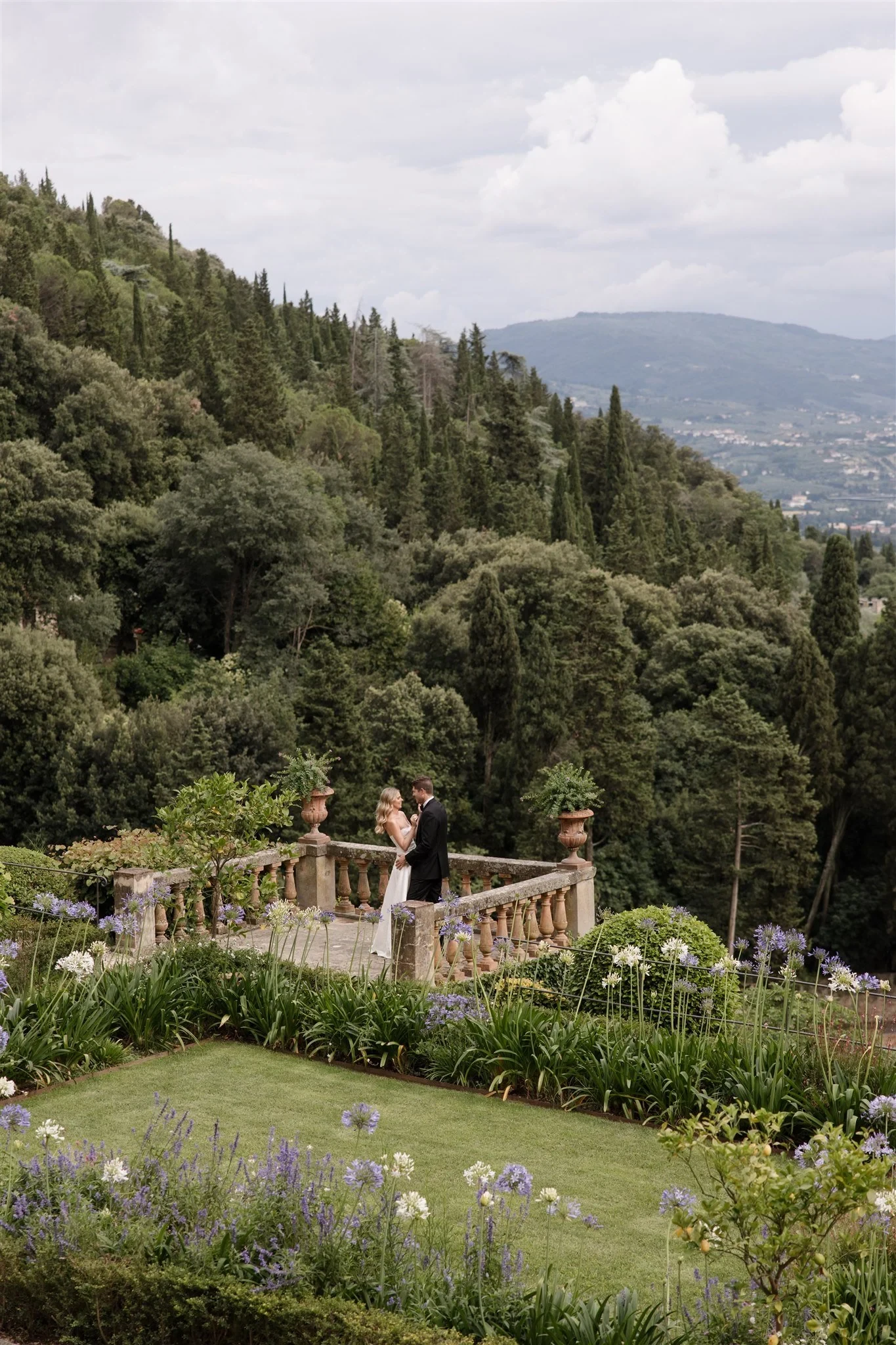 Holly and Matt standing at a stone balustrade amid the villa gardens and wooded hills, at Belmond Villa San Michele in Fiesole, overlooking Florence, Tuscany, Italy, for an Elopement Collective elopement. Photo by Khara from House of Lucie.