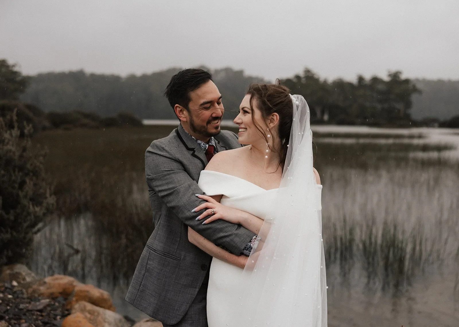 Jono embracing Sarah from behind beside lakeside reeds, with her off-the-shoulder white dress visible, at Pumphouse Point, Lake St Clair, Tasmania, for an Elopement Collective elopement. Photo by Bulb Creative.