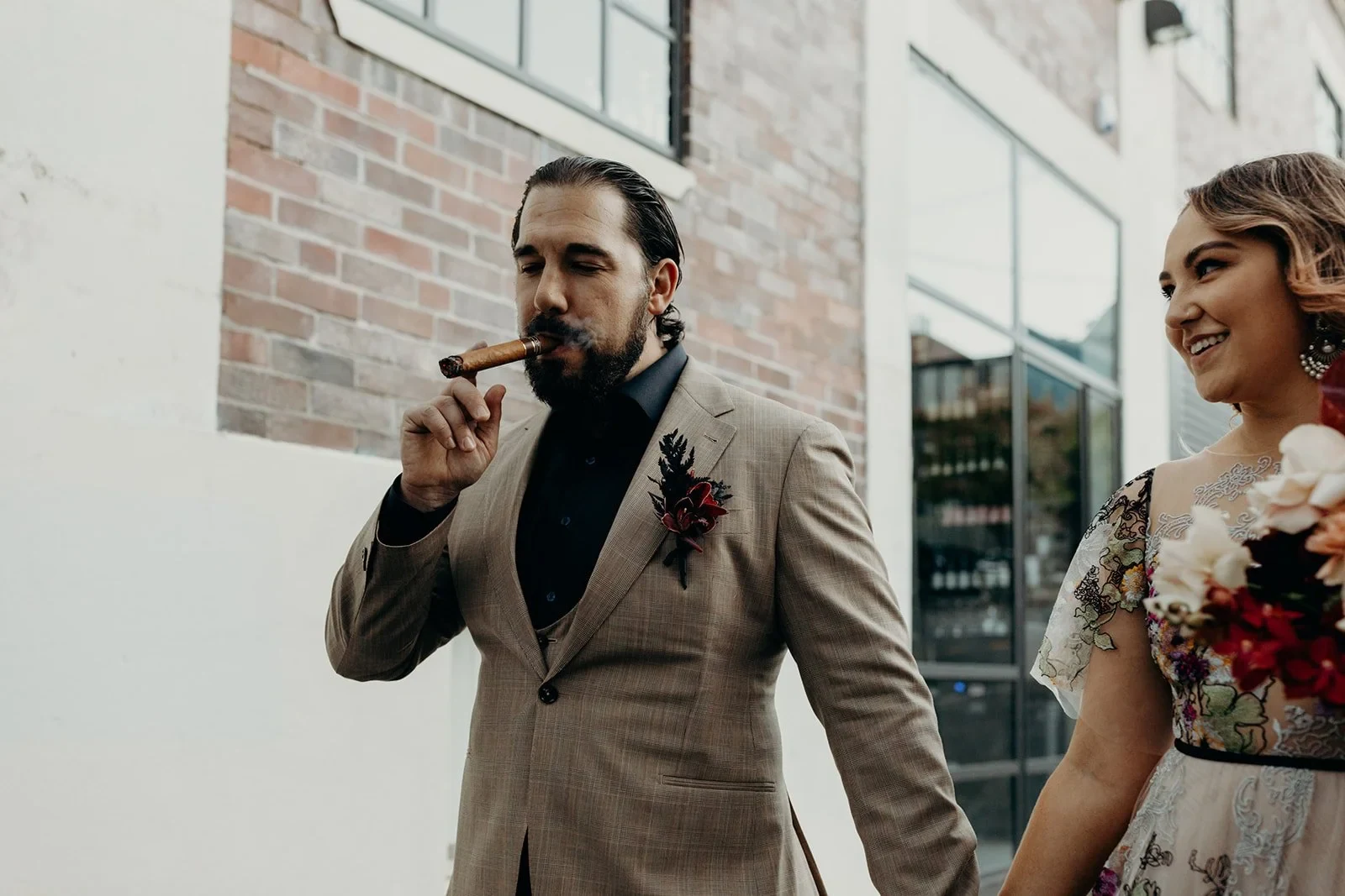 Daniel smoking a cigar beside Victoria and her bouquet by Mrs Gibbons Flowers, in Fortitude Valley, Brisbane, for an Elopement Collective elopement. Photo by Tessa Shannon.