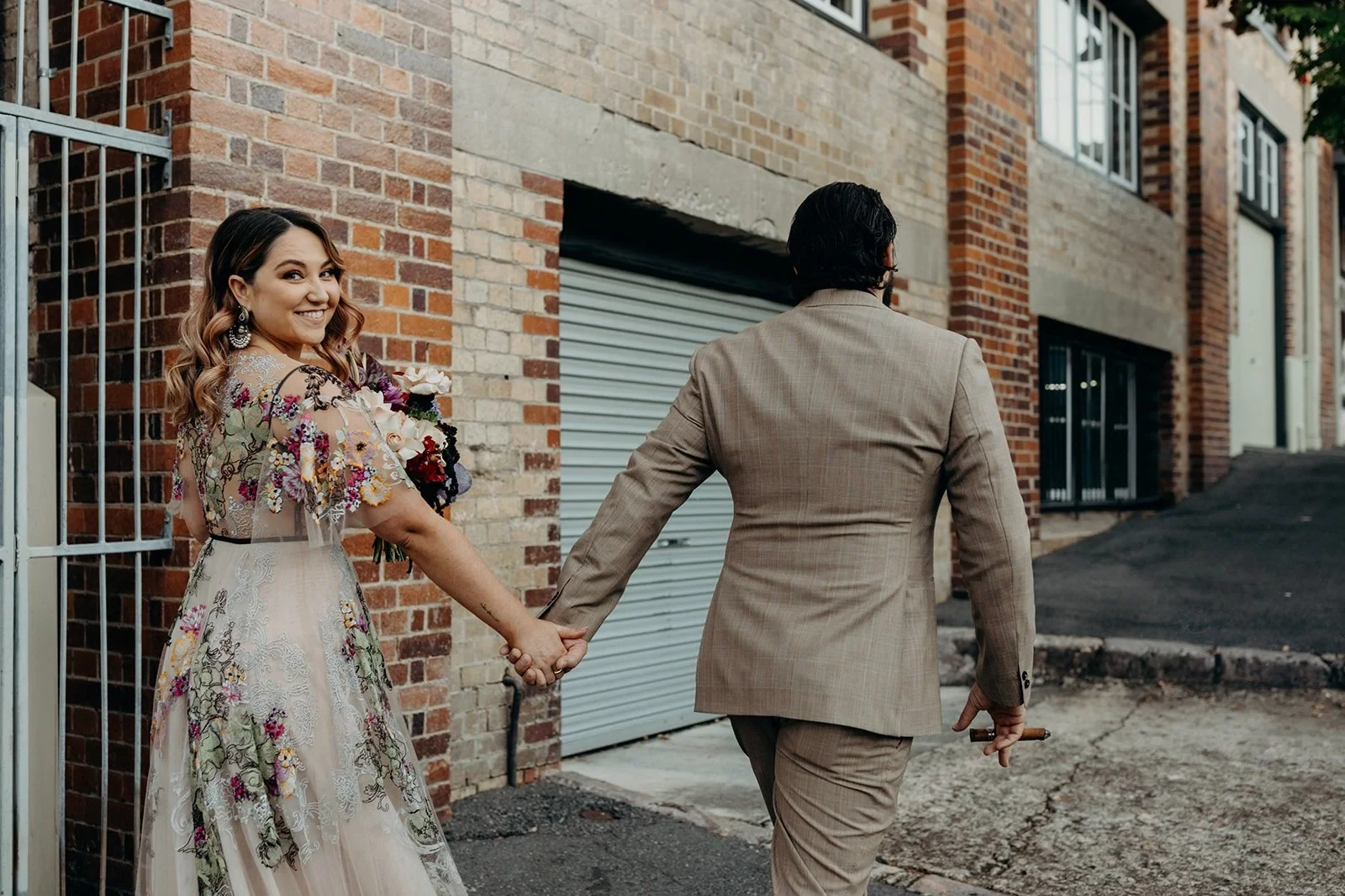 Victoria looking back over her shoulder as she and Daniel walk hand in hand along a brick-lined street, in Fortitude Valley, Brisbane, for an Elopement Collective elopement. Photo by Tessa Shannon.