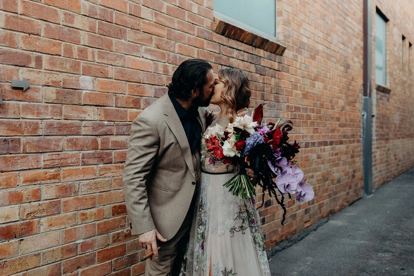 Victoria and Daniel kissing beside a red brick wall, with her bouquet by Mrs Gibbons Flowers, in Fortitude Valley, Brisbane, for an Elopement Collective elopement. Photo by Tessa Shannon.
