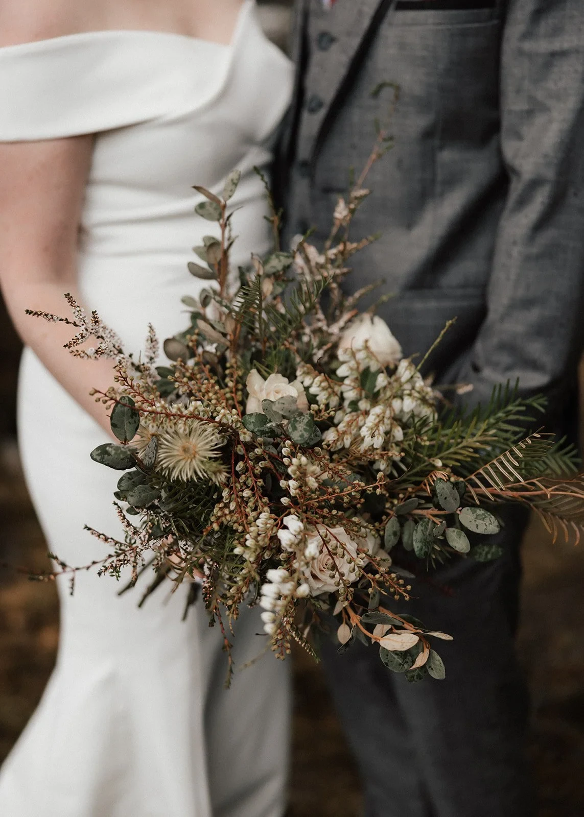 Sarah holding her bouquet by The Romantics against her white dress beside Jono’s grey suit, at Pumphouse Point, Lake St Clair, Tasmania, for an Elopement Collective elopement. Photo by Bulb Creative.