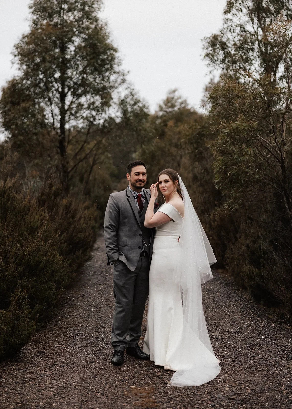 Sarah and Jono standing together on a gravel path framed by tall forest trees, at Pumphouse Point, Lake St Clair, Tasmania, for an Elopement Collective elopement. Photo by Bulb Creative.