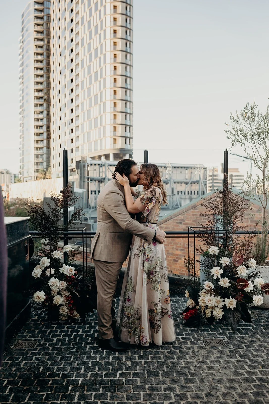 Victoria and Daniel kissing beside white ceremony flowers by Mrs Gibbons Flowers on the rooftop terrace, at Agnes Restaurant in Fortitude Valley, Brisbane, for an Elopement Collective elopement. Photo by Tessa Shannon.