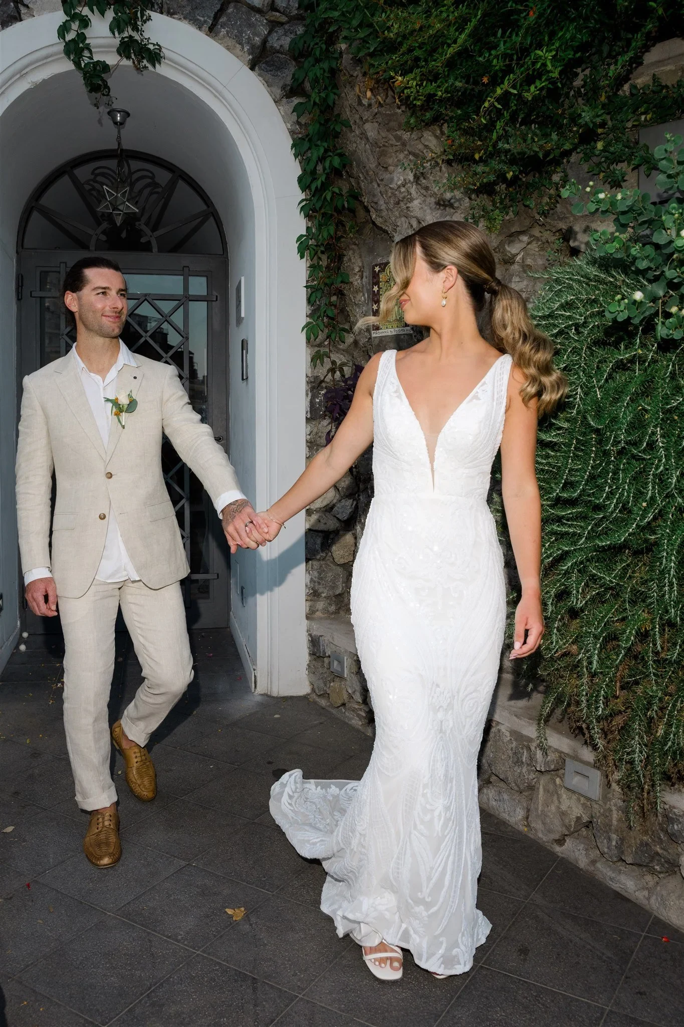 Sarah and Josh stepping through an arched stone doorway holding hands, her white dress trailing behind her, in Positano on Italy’s Amalfi Coast, for an Elopement Collective elopement. Photo by James from House of Love.